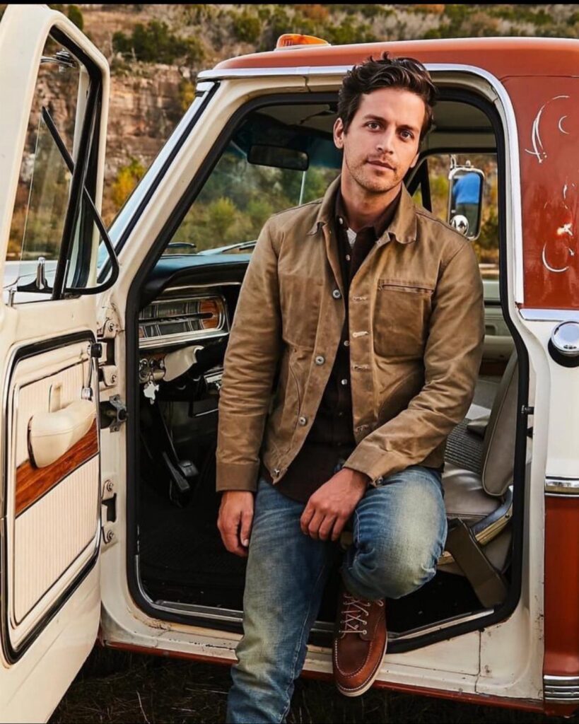 A man in a tan utility jacket and blue jeans stepping out of a vintage truck.