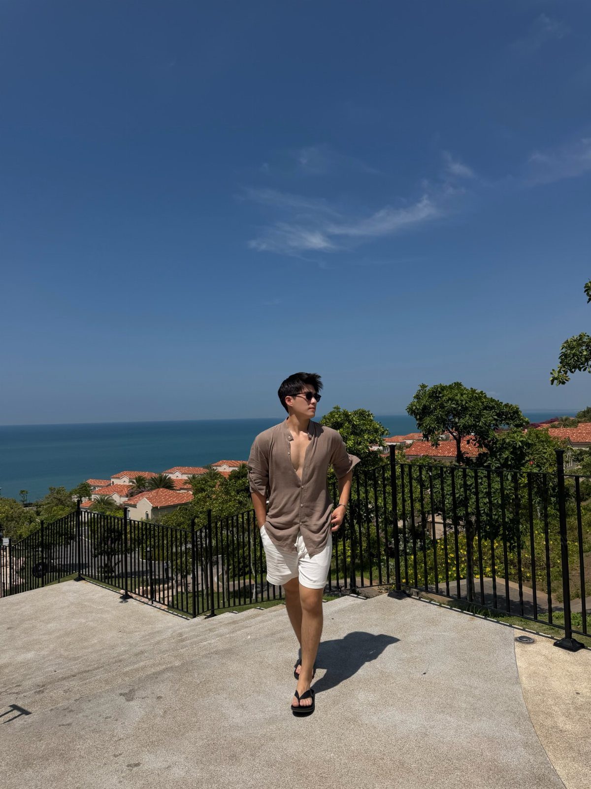 A man in a brown shirt and white shorts standing by a fence overlooking the sea.
