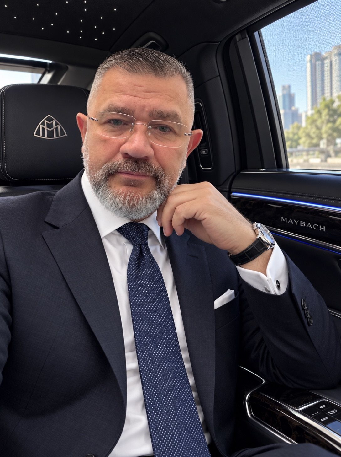 A bearded man in a navy suit and tie sitting inside a luxury Maybach vehicle.