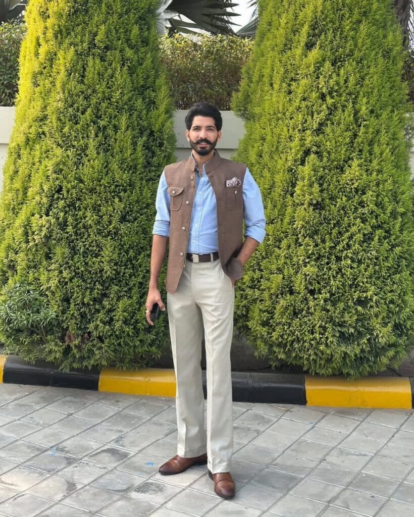 Man wearing a brown waistcoat, light blue shirt, and cream tailored trousers standing outdoors.