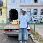 Man in white t-shirt and denim shorts standing in front of a vintage tan car.