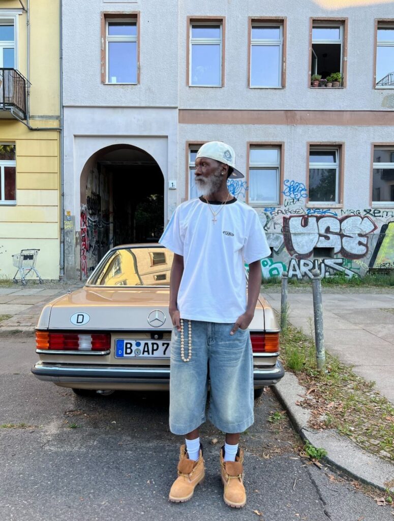 Man in white t-shirt and denim shorts standing in front of a vintage tan car.