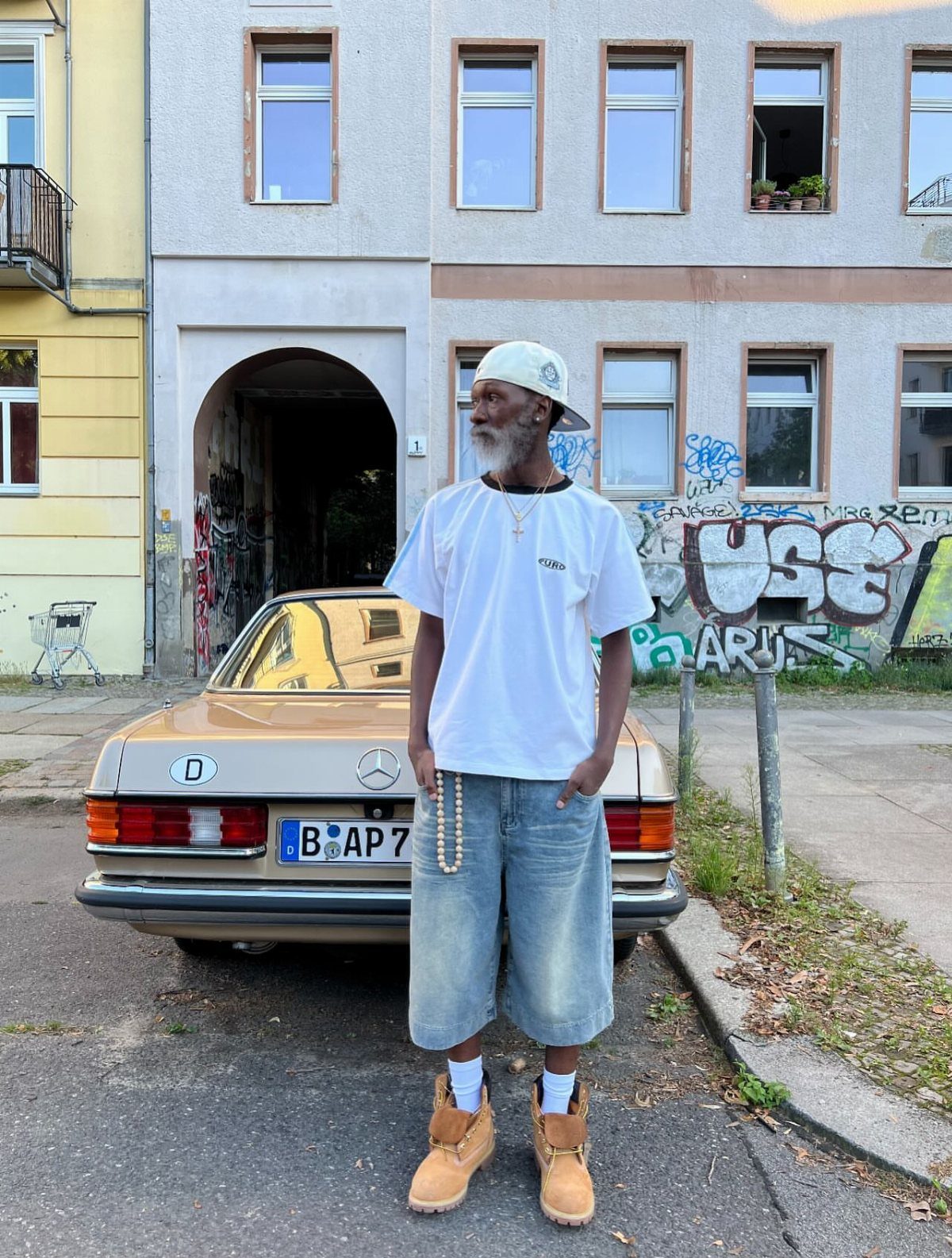 Man in white t-shirt and denim shorts standing in front of a vintage tan car.
