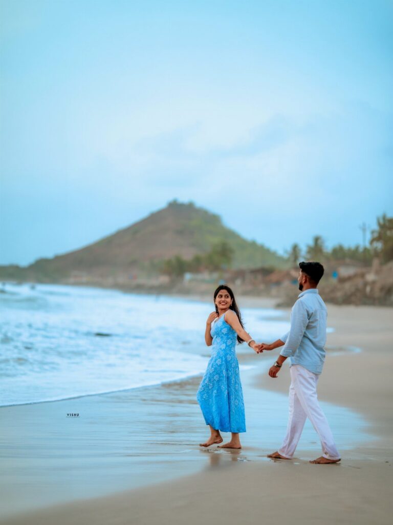 A man in a light blue shirt and white pants holds hands with a woman in a blue dress on a beach.