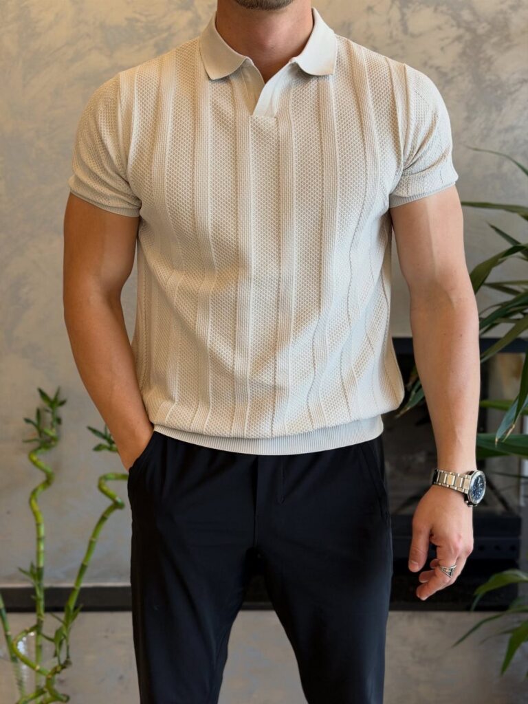 A man wearing a textured cream polo shirt and black trousers standing near green plants.