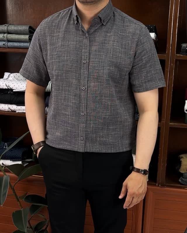 Man wearing a grey short-sleeved button-down shirt and black dress pants standing indoors.