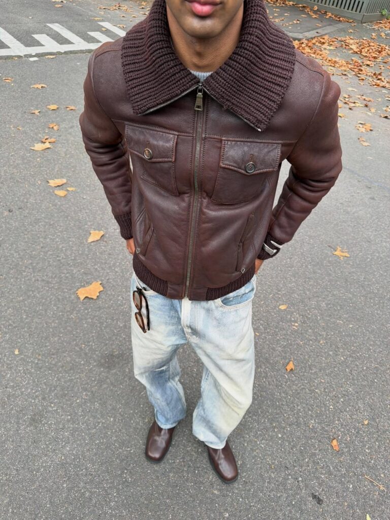 Man wearing a brown leather jacket with a knit collar, light denim jeans, and brown shoes.