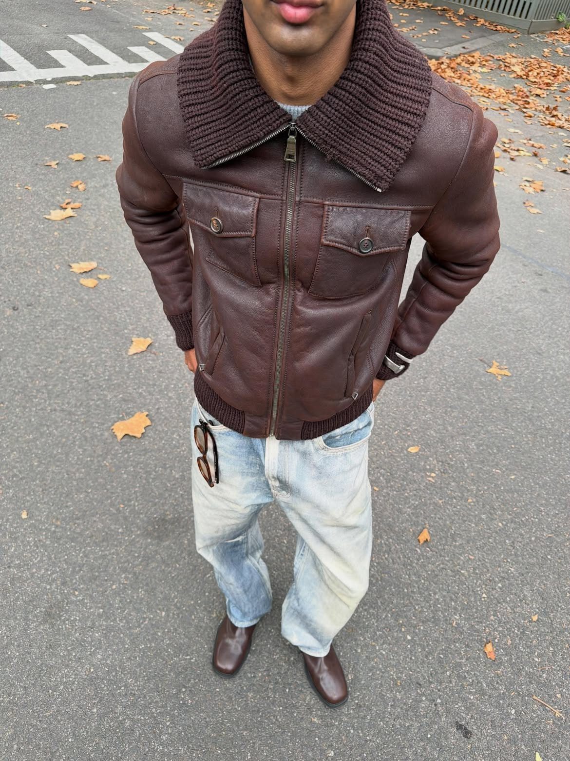 Man wearing a brown leather jacket with a knit collar, light denim jeans, and brown shoes.