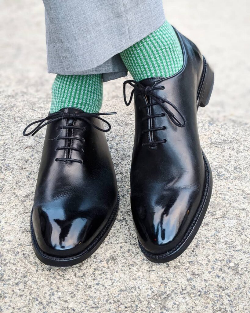 Black leather oxford shoes worn with green socks and grey trousers on concrete.