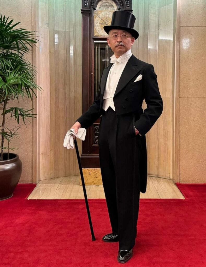 Man in formal white tie attire with a top hat and cane standing on a red carpet.