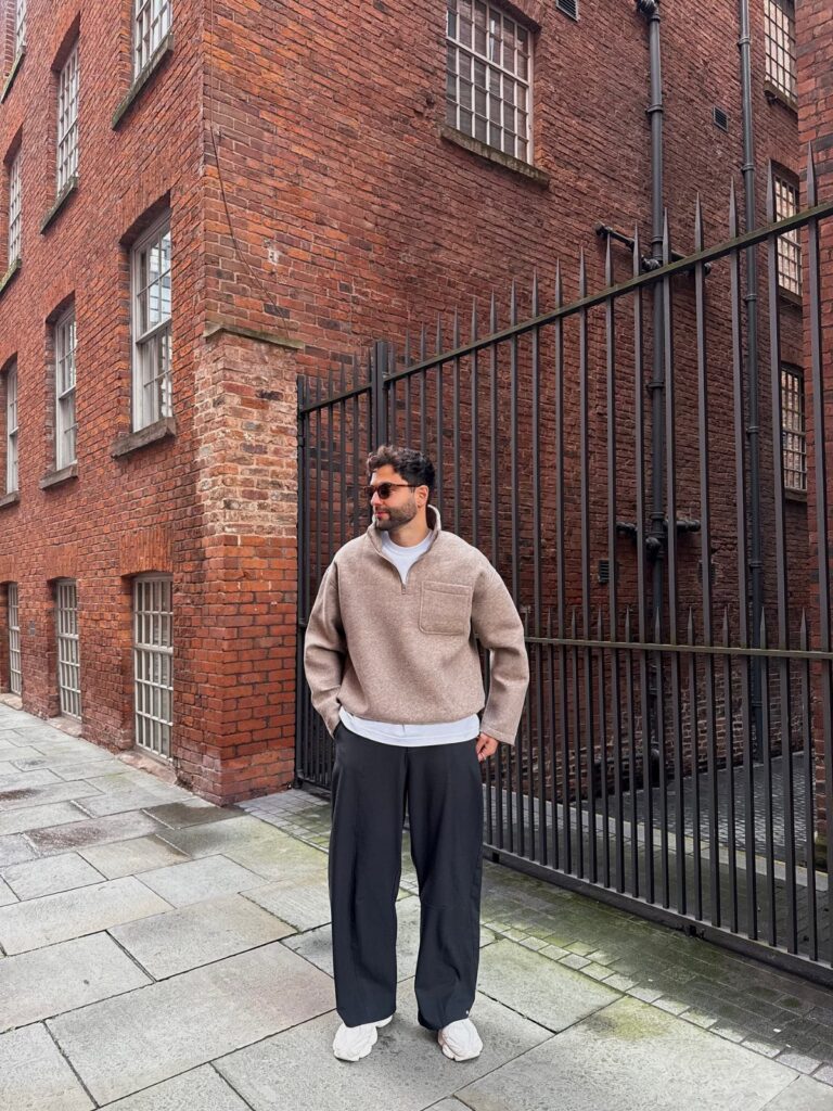 A man wearing a beige fleece and black wide-leg trousers standing by a brick building.