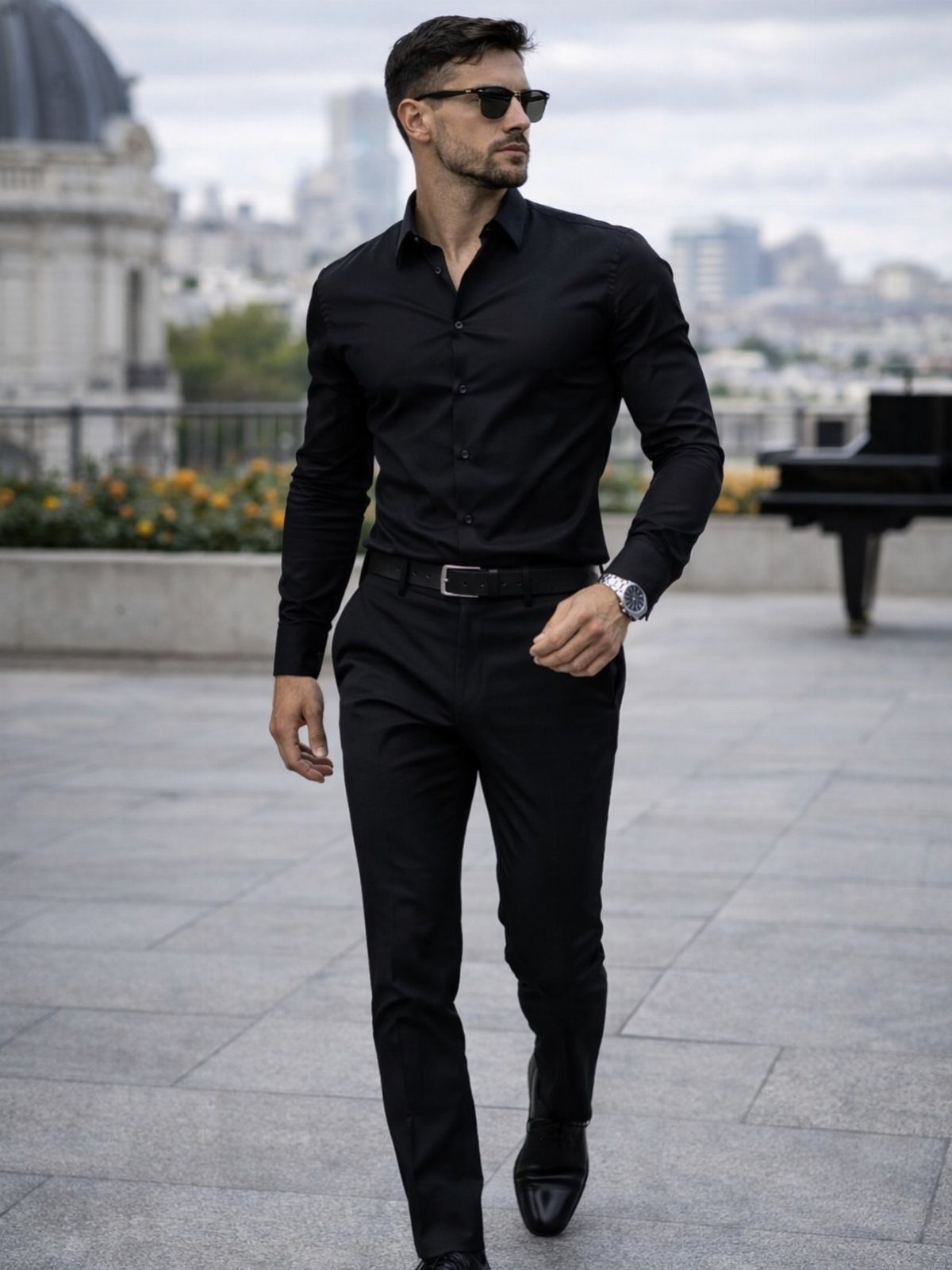 A man in an all-black formal outfit walking on a city terrace wearing sunglasses.