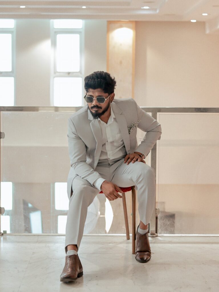 A man in a light grey suit and sunglasses sitting on a chair indoors.