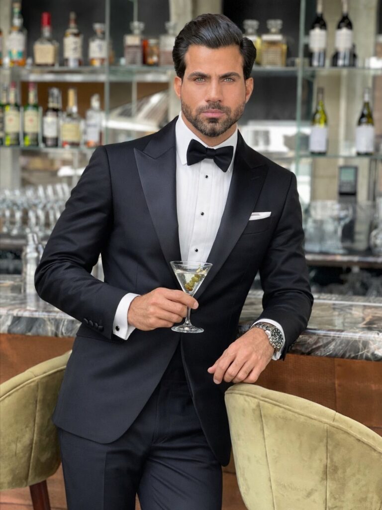 A man in a black tuxedo and bow tie holding a martini glass at a bar.