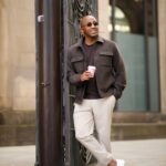 A man in a brown wool shacket and beige trousers leans against a pillar holding coffee.