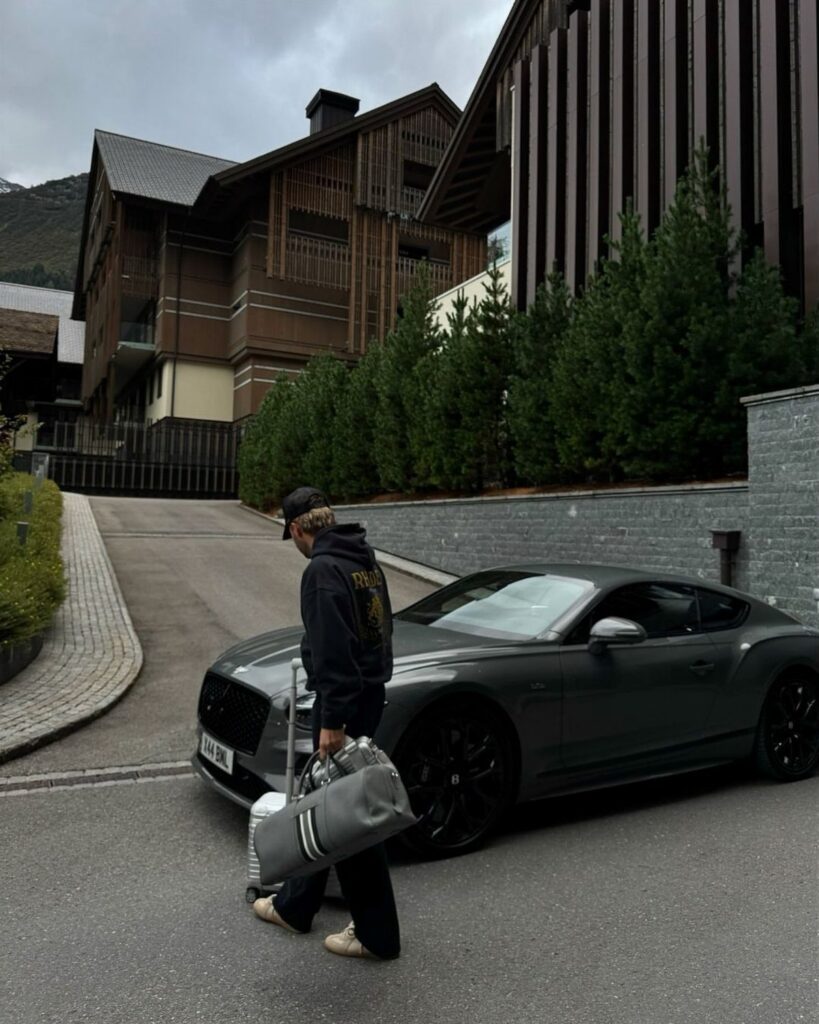 A person walking past a matte grey luxury sports car parked near modern wooden mountain chalets.