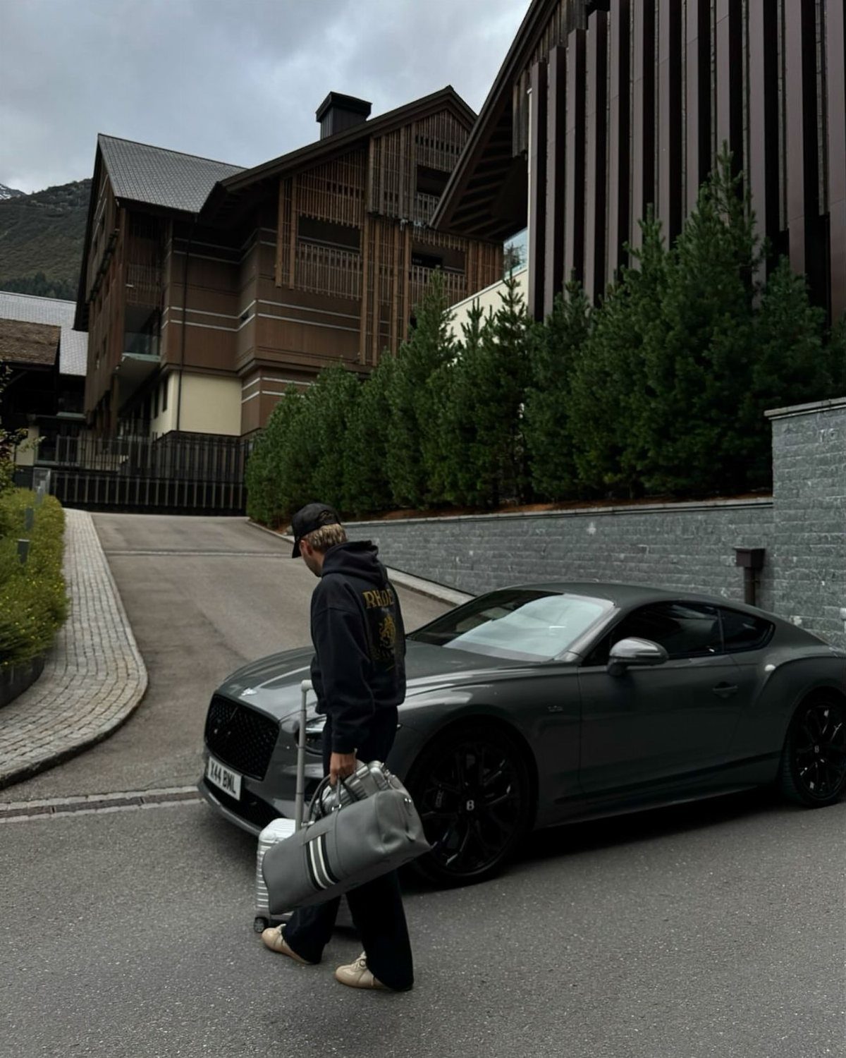 A person walking past a matte grey luxury sports car parked near modern wooden mountain chalets.