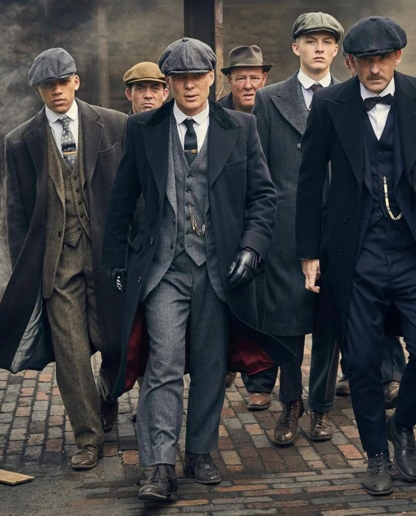 Men walking in a group wearing vintage Peaky Blinders style three-piece suits and flat caps.