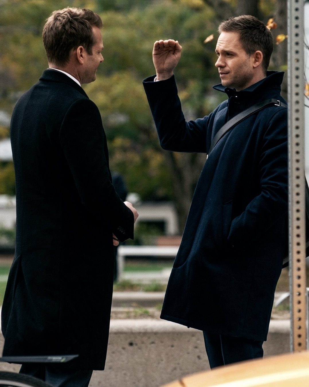 Two men wearing sophisticated dark overcoats talking outdoors near a street.