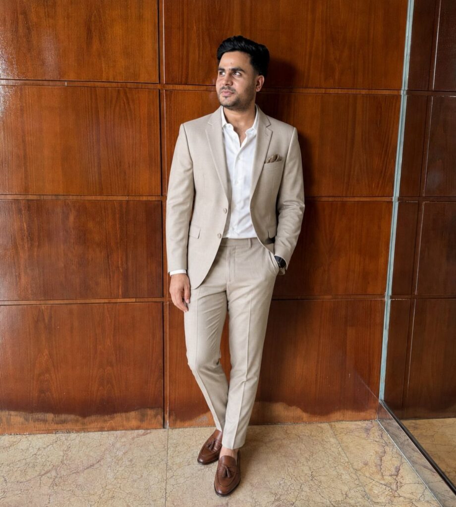 A man in a tan suit and white dress shirt posing against a wooden wall.