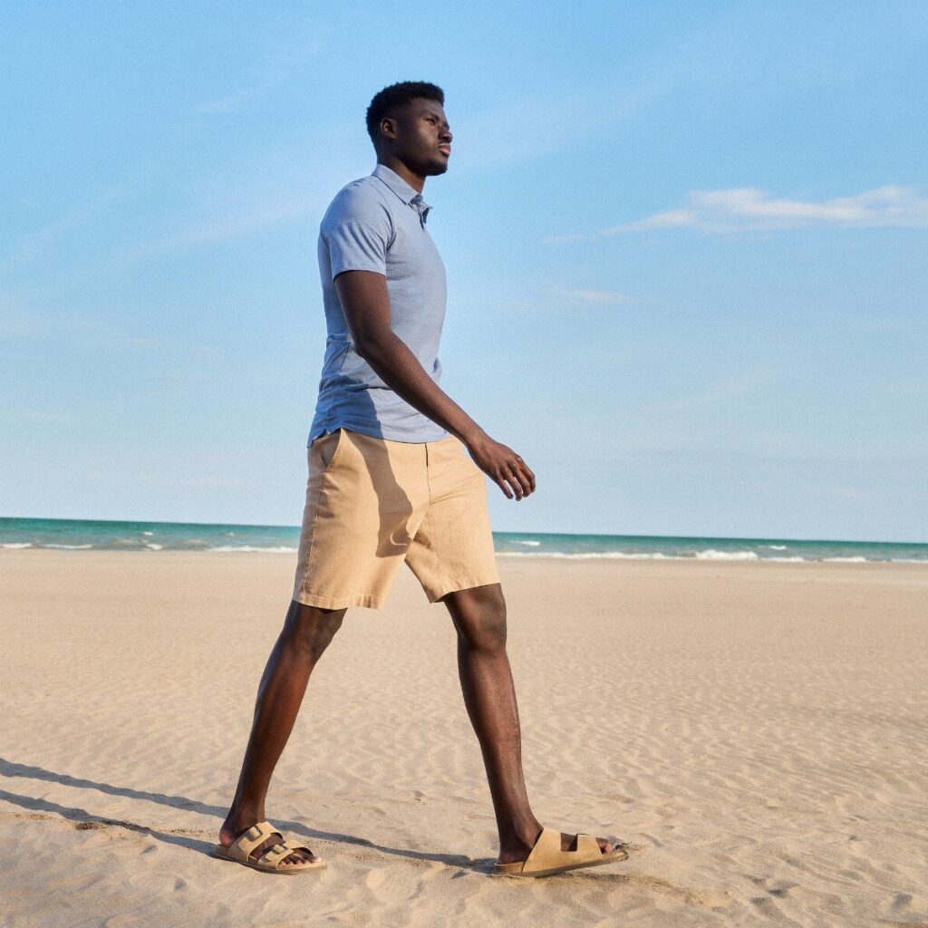 A tall man in a blue polo and tan shorts walking along a sunny beach.