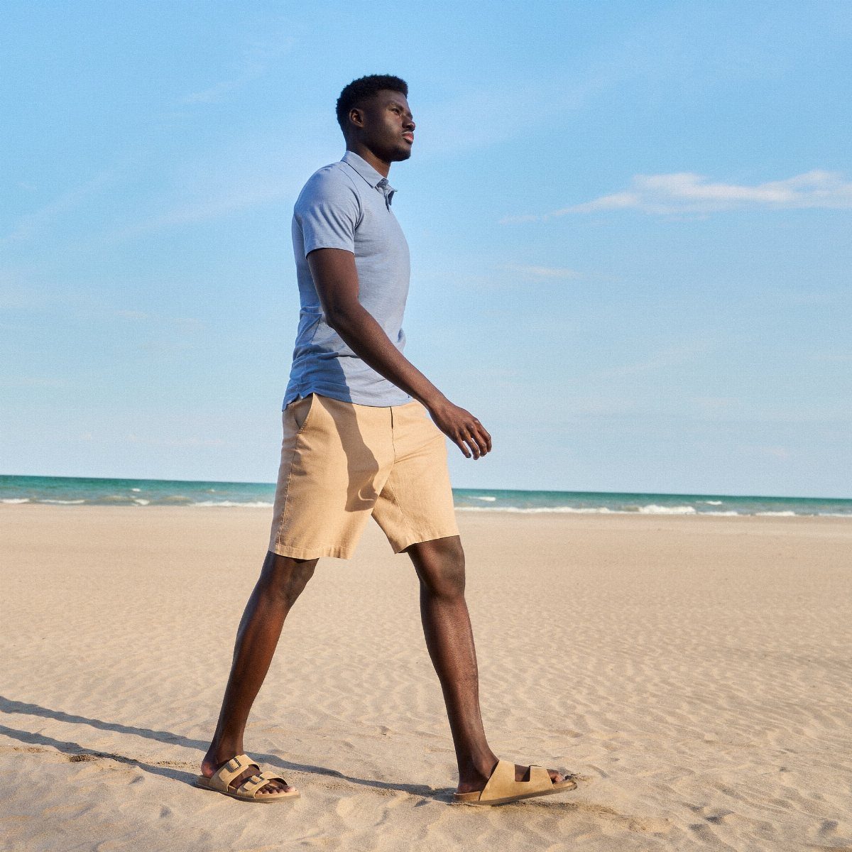 A tall man in a blue polo and tan shorts walking along a sunny beach.