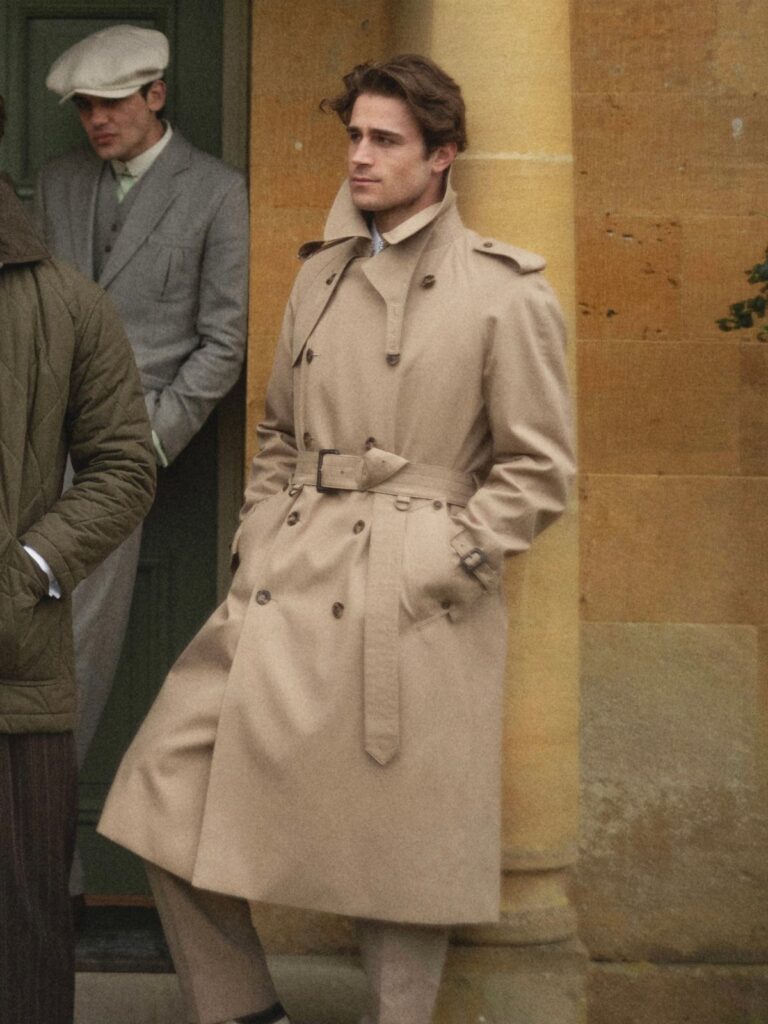 A man wearing a classic tan double-breasted trench coat standing against a stone wall.