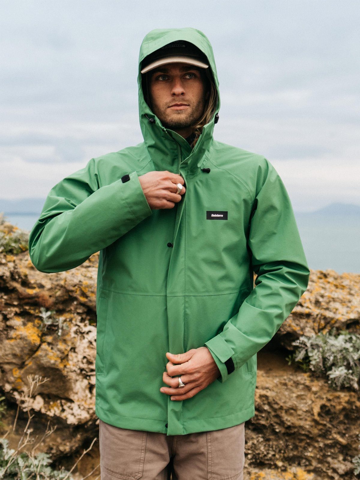 A man wearing a green hooded waterproof jacket standing in a rocky outdoor landscape.